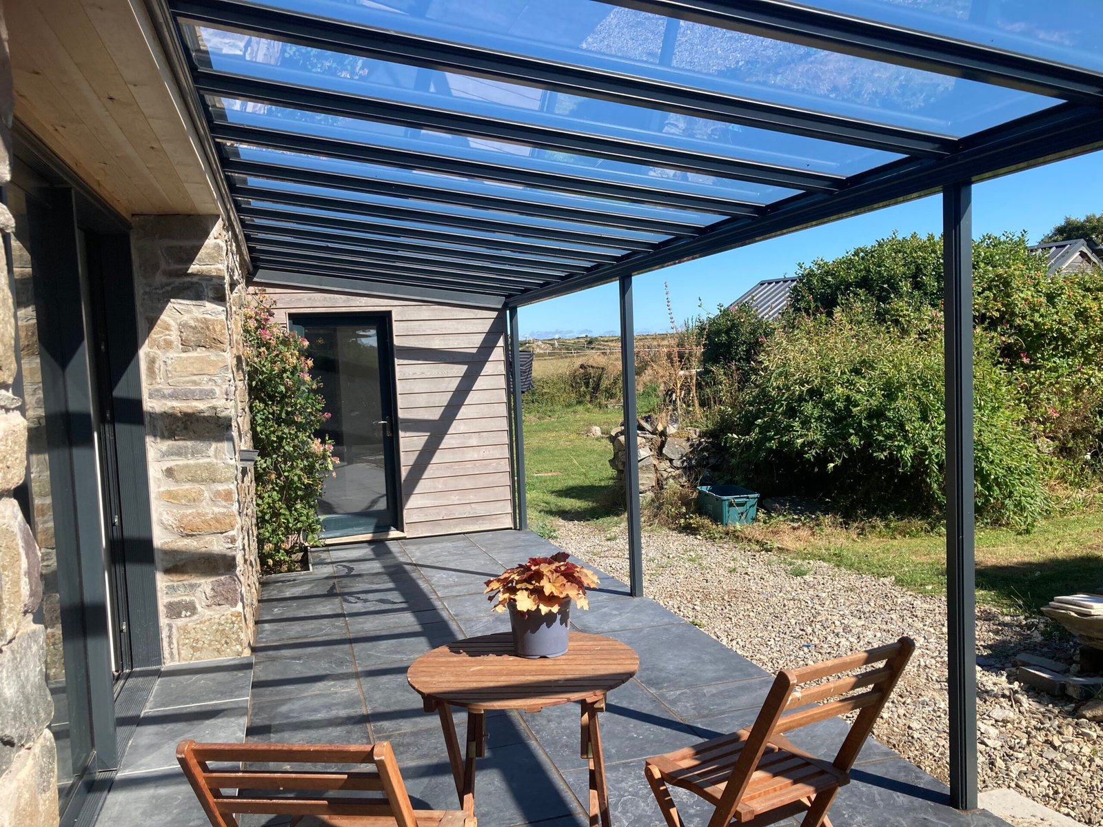 Outdoor glass-roofed patio with stone and wood walls, black metal framing, and wooden seating near a garden.